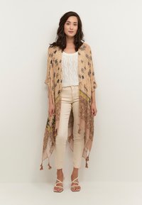 Beige mustriga kimono, mille- ja loomamustriga aktsentidega, paeltega allservas, kantud valge pitsist pluusi ja kreemivärvi pükste peal.