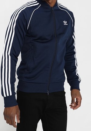 Homme portant une veste de survêtement Adidas bleu marine avec des détails à trois bandes blanches sur les manches et une fermeture éclair devant, associée à un pantalon noir.