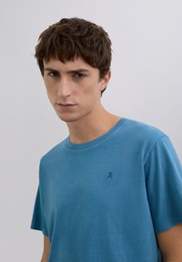 Ung mand med kort krøllet brunt hår iført en blå t-shirt med et lille mørkt kraniumlogo på brystet mod en ensfarvet lys baggrund.