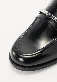 Mocassino in pelle nera con finitura lucida, caratterizzato da dettagli cuciti e una fascia in metallo argentato attraversa la parte anteriore.