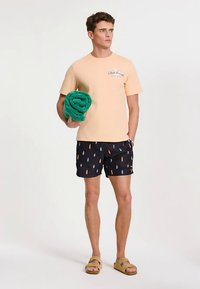 Őszibarackszínű póló logóval, fekete fürdőnadrág színes, függőleges mintákkal, és egy zöld törölköző. Szandál van a lábakon.