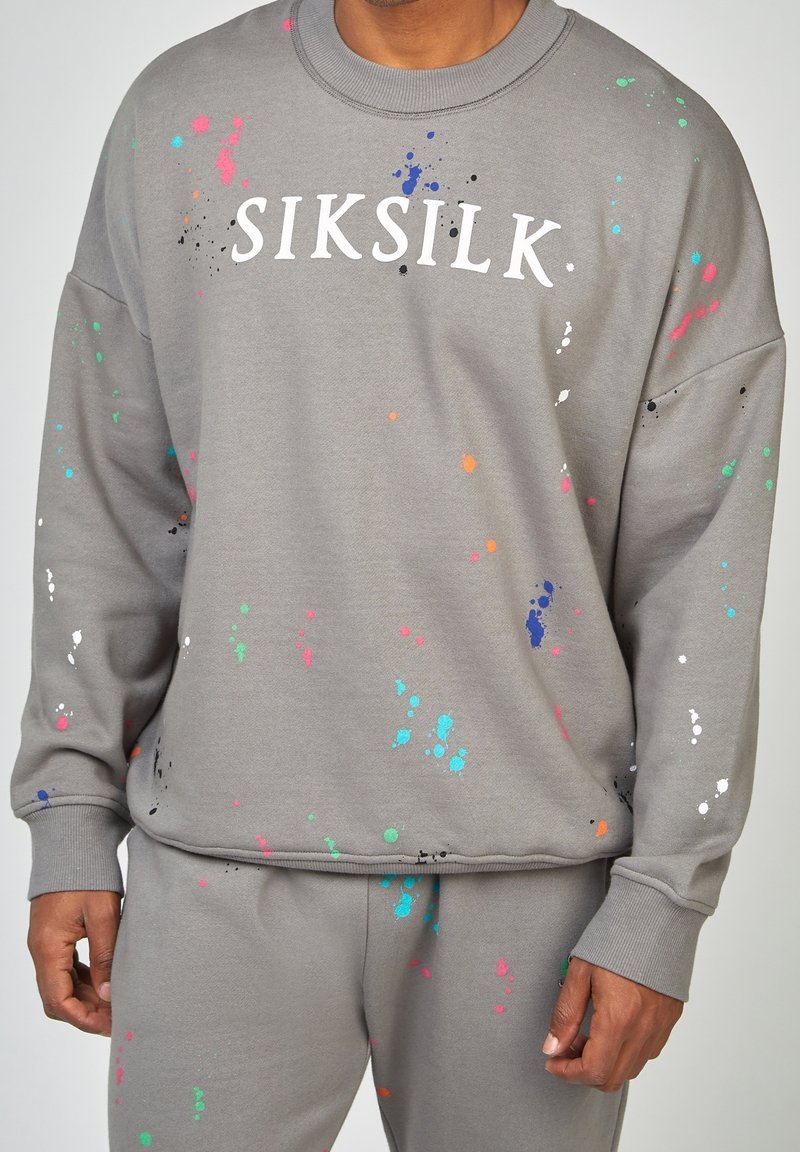 Sudadera gris con manchas de pintura multicolores, con cuello redondo y corte relajado. El logo de la marca "SIKSILK" se muestra de manera prominente en blanco.