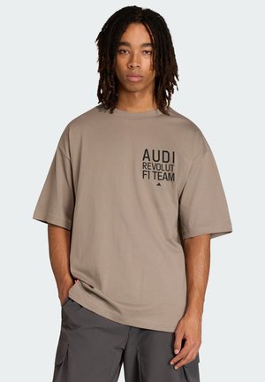 Giovane con dreadlocks di media lunghezza che indossa una maglietta beige larga con la scritta "AUDI REVOLUT FIT TEAM" e pantaloni cargo scuri.