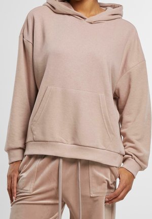 Person iført matchende beige hættetrøje med frontlomme og fløjlsbukser med snørre og sidelommer, stående mod en ensfarvet baggrund.