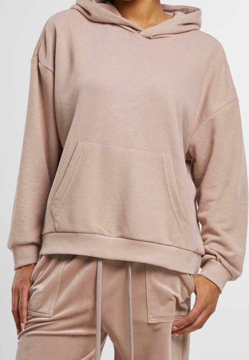 Personne portant un sweat à capuche beige assorti avec poche avant et pantalon en velours à cordon avec poches latérales, debout devant un fond uni.