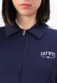 Navy blue zip-up sweatshirt with a folded collar, featuring a white "GAP NYC" logo and a silver zipper. Soft fabric with a smooth texture.
