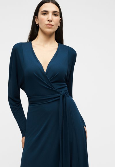Γυναίκα με μακριά σκούρα μαλλιά που φοράει βαθιά μπλε φόρεμα τύπου wrap με μακριά μανίκια και δεμένη μέση σε απλό φωτεινό φόντο.