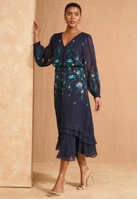 Rochie bleumarin cu mâneci lungi și transparente, decolteu în V, broderie florală, volane în straturi la tiv, asortată cu sandale cu toc aurii.