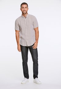 Camicia a maniche corte, color beige chiaro, in tessuto texture, abbinata a jeans scuri e sneakers bianche, in piedi contro uno sfondo neutro.