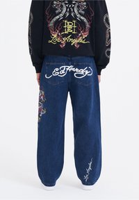Baggy blå denimjeans med vita broderade "Ed Hardy" och "Los Angeles" logotyper, samt färgglada drake-grafik på sidan.