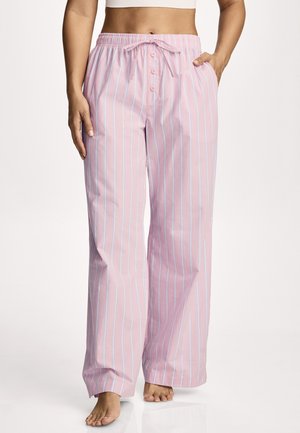 Woman wearing loose pink pajama pants with white vertical stripes, elastic waistband, button fly, and drawstring, standing barefoot.