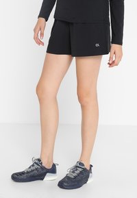 Person wearing black athletic shorts with white logo, black long-sleeve top, and navy blue sneakers with white soles and laces.