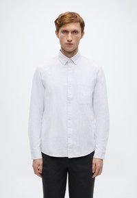 Λευκό πουκάμισο με κουμπιά και κλασικό γιακά, κάθετη ριγέ υφή και τσέπη στο στήθος. Φορεμένο με σκούρο παντελόνι, στέκεται μπροστά σε απλό φόντο.