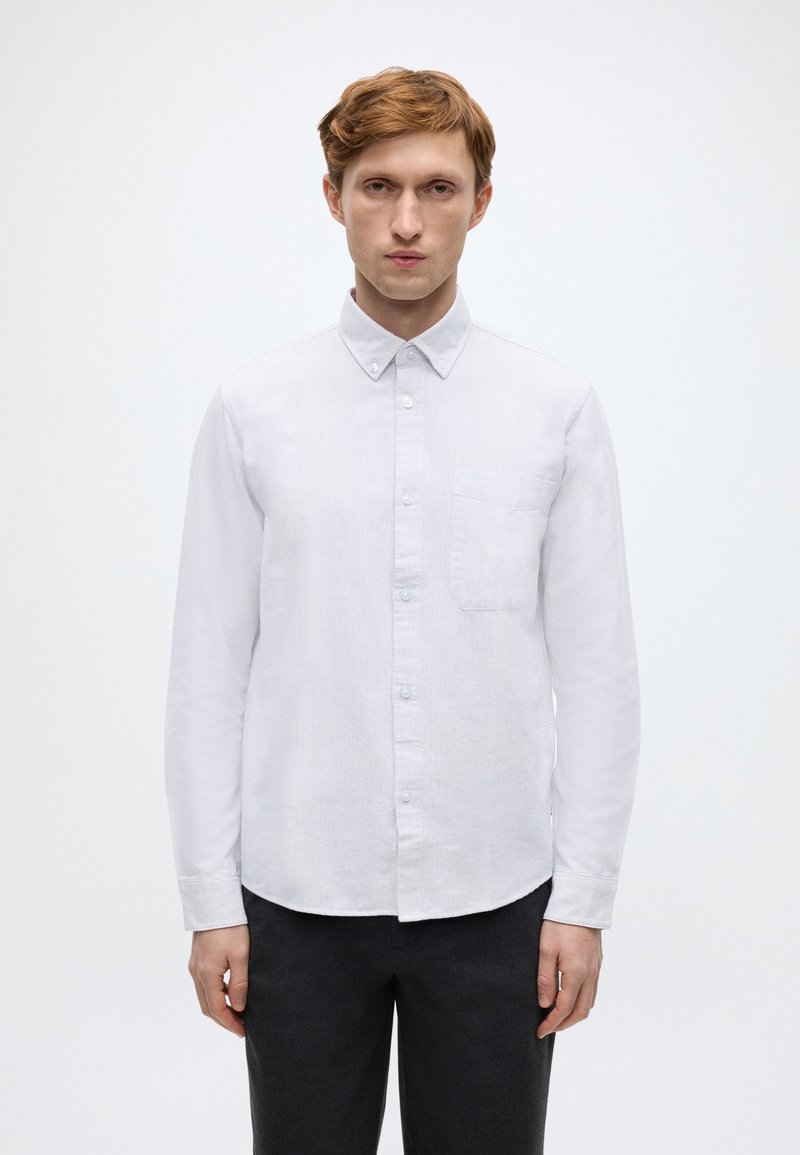 Λευκό πουκάμισο με κουμπιά και κλασικό γιακά, κάθετη ριγέ υφή και τσέπη στο στήθος. Φορεμένο με σκούρο παντελόνι, στέκεται μπροστά σε απλό φόντο.