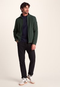 Dark green fleece jacket with a high collar, zipped front, paired with a navy sweater, black pants, and white sneakers with black stripes.