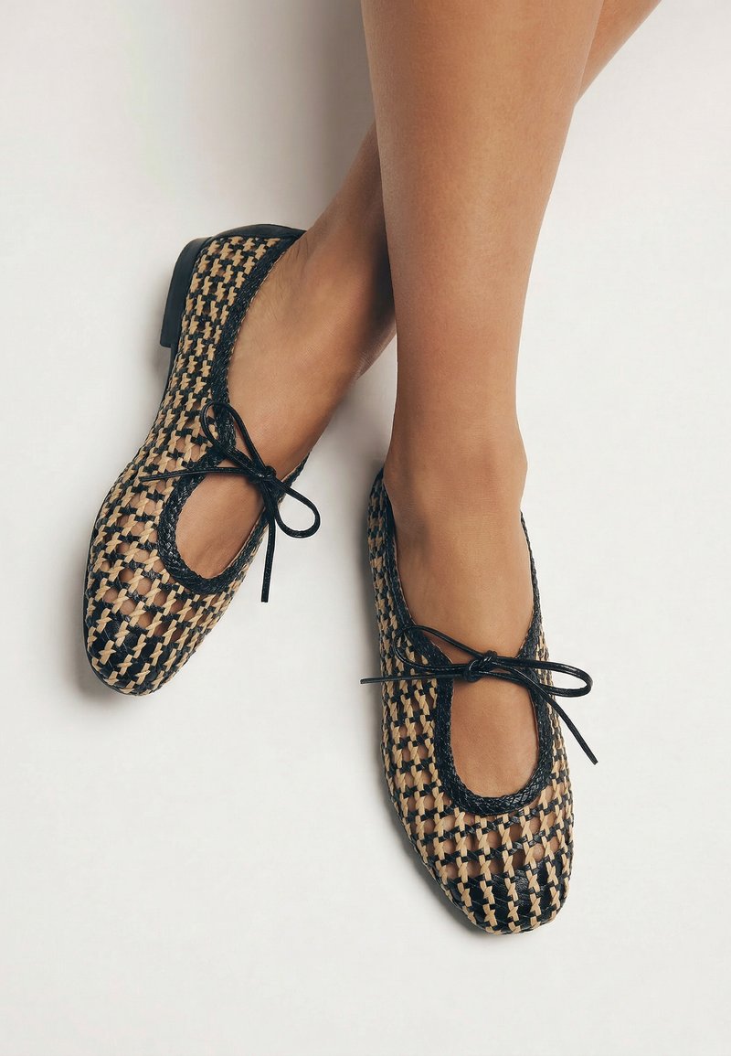 Pies con zapatillas de ballet tejidas en negro y beige con ribete y lazos negros, cruzados sobre un fondo blanco liso.