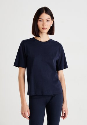 Donna con capelli lisci e scuri, che indossa una maglietta a maniche corte blu scuro e pantaloni abbbinati, in piedi su uno sfondo chiaro e neutro.