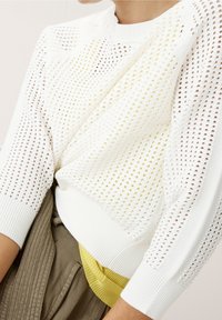 Biały dziany sweter z teksturowanym wzorem, żółtymi akcentami na dole oraz ściągaczami na mankietach i kołnierzu; noszony na beżowej spódnicy.