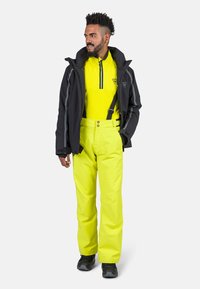 Completo da sci con giacca nera impermeabile, pantaloni gialli vivaci e una maglia gialla a maniche lunghe con zip a quarti. Tessuto testurizzato e numerose tasche.