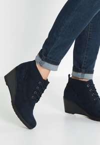 Bottines compensées en daim bleu marine avec design lacé et œillets métalliques. Fini texturé et bout arrondi, associées à un jean à revers.
