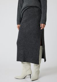 Jupon tricoté gris charbon avec fente sur le côté, associé à des bottes à talons hauts crème. Il dispose d'une taille ajustée et d'un tissu doux et texturé.