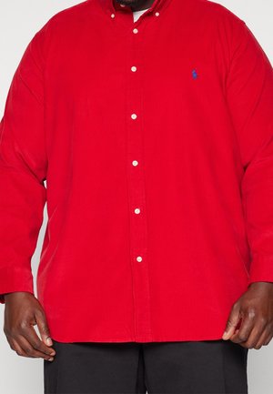 Mężczyzna ubrany w jasnoczerwoną, zapinaną na guziki koszulę z długim rękawem, z małym niebieskim haftowanym logo na lewej piersi, w parze z czarnymi spodniami.