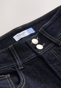 Lainelõikega kõrge vöökohaga teksapüksid tumesinises pesus, kuldsete detailide ja kahe nupu kinnitusega ning kontrastse pruunika õmblusega. Silt on nähtav sees.