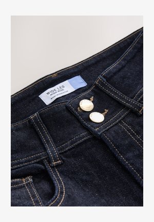 Vaqueros de pierna ancha y talle alto en lavado oscuro con herrajes dorados, cierre de dos botones y costuras contrastantes en color marrón claro. Etiqueta visible en el interior.