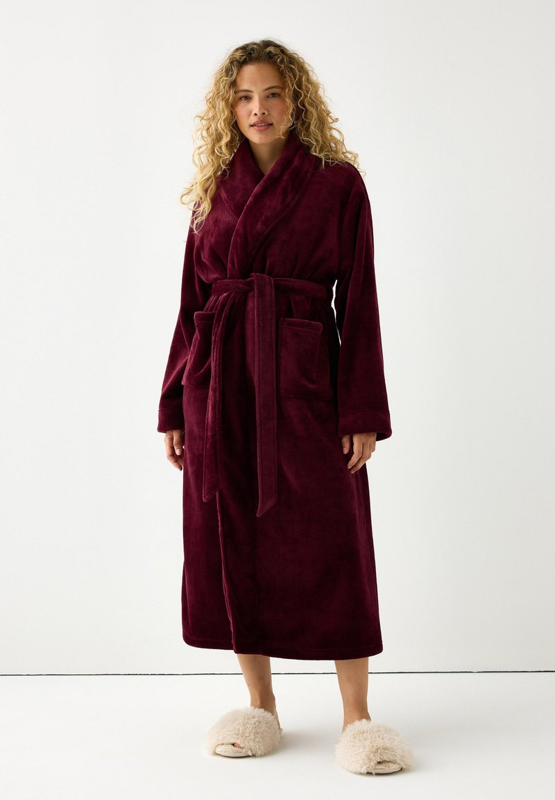 Burgundy plush kylpytakki, jossa on vyö, pitkät hihat ja kaksi etutaskua, yhdistettynä beigekakkaroisiin tossuihin valkoisella taustalla.