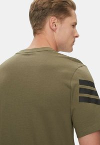 T-shirt verde oliva a maniche corte con scollatura rotonda e tre strisce nere sulla spalla sinistra. Tessuto morbido.