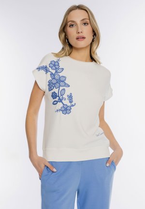 Kobieta w białej bluzce z krótkim rękawem, z niebieską kwiatową haftem na ramieniu i jasnoniebieskich spodniach, z rękami w kieszeniach.