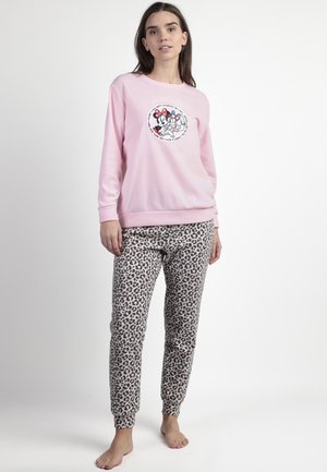 Mujer de pie descalza, vistiendo un sudadera rosa con personajes de dibujos animados y pantalones de pijama con estampado de leopardo, contra un fondo liso.