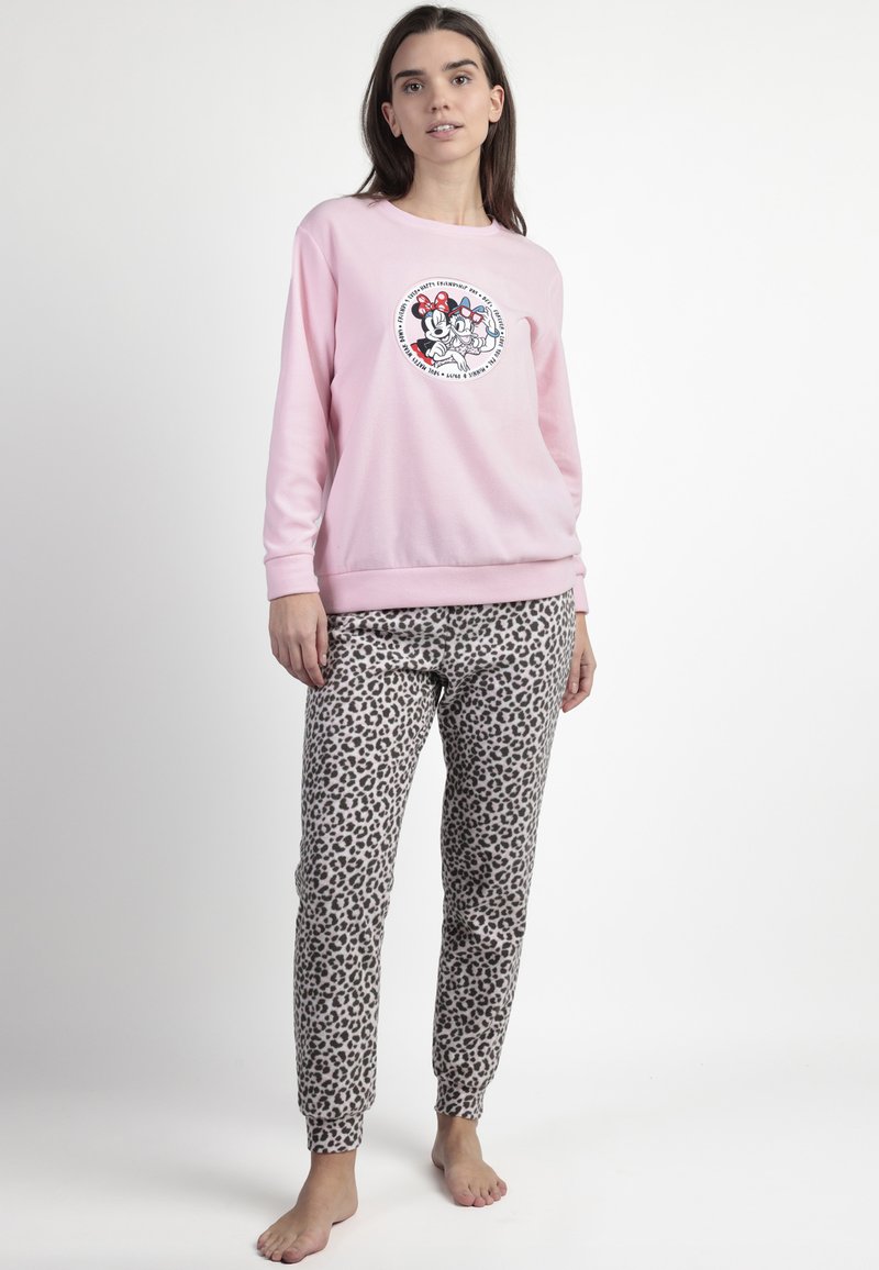 Mujer de pie descalza, vistiendo un sudadera rosa con personajes de dibujos animados y pantalones de pijama con estampado de leopardo, contra un fondo liso.