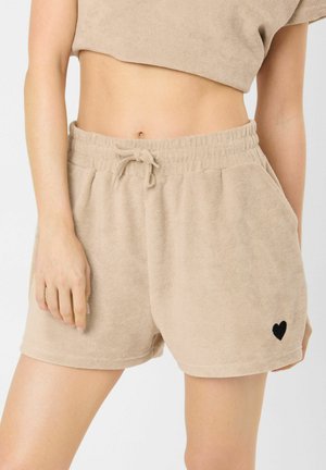 Beige terry cloth shorts with elastic waistband and drawstring, featuring a small black heart on the lower left leg, worn with matching cropped top.
