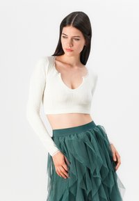Hvit croppet topp med lange ermer, med bølget hals og teksturert mønster, kombinert med et lagdelt tulle-skjørt i teal.