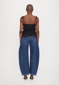 Person, die ein schwarzes ärmelloses Oberteil und weite blaue Jeans trägt, barfuß vor einem weißen Hintergrund steht und von der Betrachterin abgewandt ist.