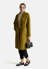 Cappotto di lana verde oliva con ampio colletto, due tasche frontali e bottoni di legno. Abbinato a pantaloni neri cropped e scarpe, tiene in mano una piccola borsa nera.