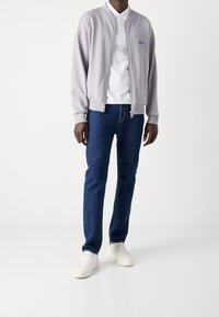 Mand iført en lys grå zip-up jakke, hvid polo skjorte, blå jeans og hvide sneakers, stående foran en ensfarvet hvid baggrund.