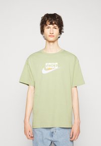 T-shirt en coton vert clair avec un logo Nike blanc et un design de texte. Coupe décontractée avec des manches courtes et un col rond.