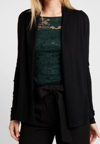 Svart cardigan med öppen front, mjukt material och långa ärmar, lager-på-lager över en mörkgrön spets-topp med blommönster och rund halsringning.