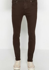 Jean slim marron foncé porté avec des baskets blanches en toile sur un fond clair uni.