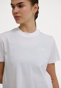 Young woman with hair in a braid wears a plain white crew neck t-shirt with a small chevron logo on the chest, looking left.