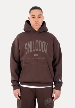 Mand iført brun hættetrøje og matchende bukser med teksten "SMILODOX Essential Sportswear" foran, stående mod en ensfarvet baggrund.