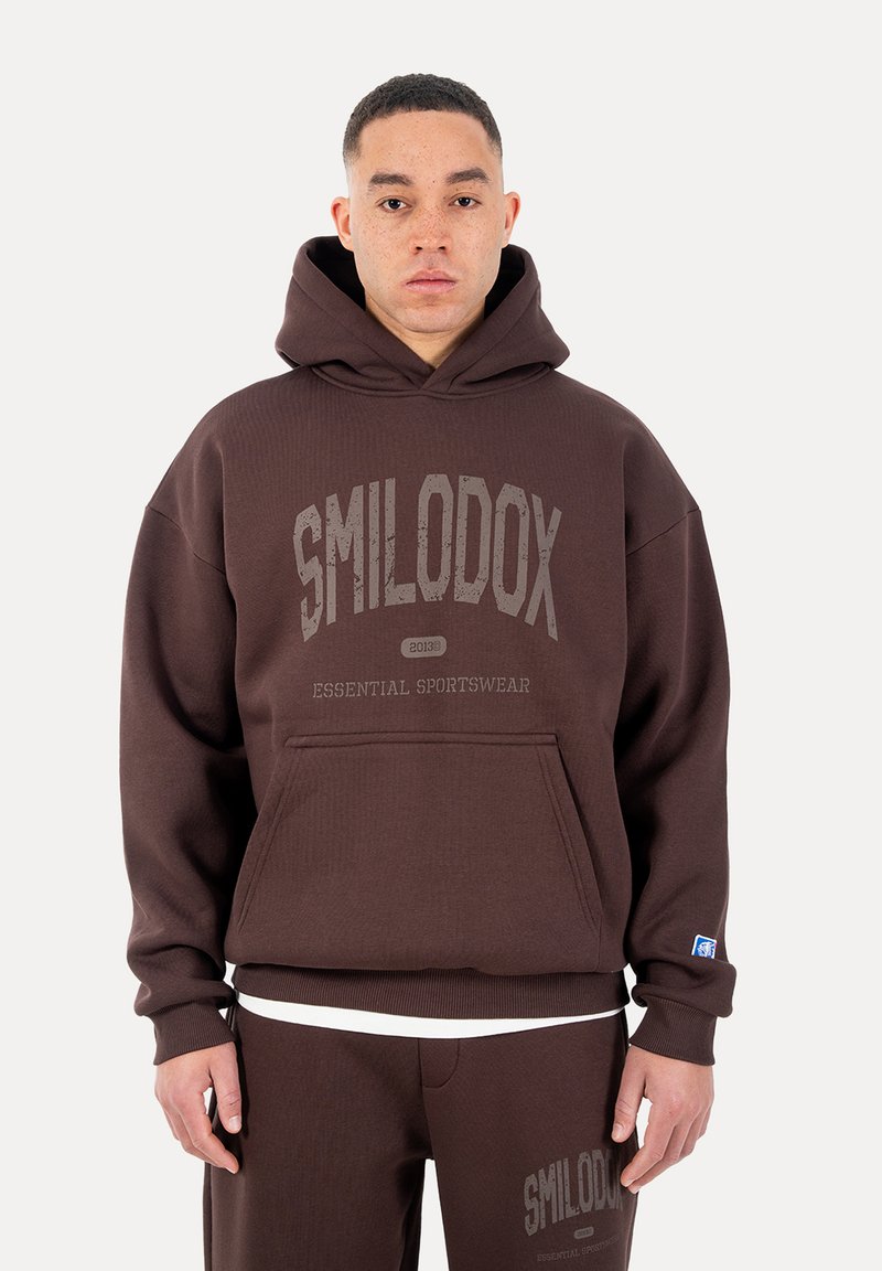 Hombre con sudadera con capucha marrón y pantalones a juego con el texto "SMILODOX Essential Sportswear" en el frente, posando contra un fondo liso.