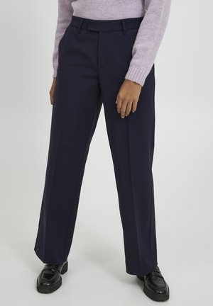 Pantalons larges bleu marine avec une texture lisse, dotés d'une taille cintrée et d'un pli avant, associés à des chaussures noires imposantes.