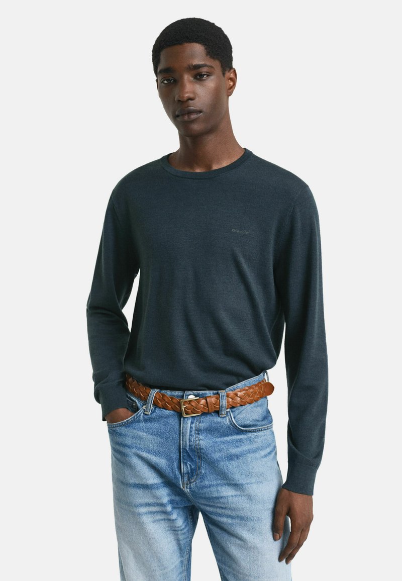 Jeune homme portant un pull à manches longues vert foncé rentré dans un jean bleu clair avec une ceinture en cuir tressé marron, une main dans la poche.