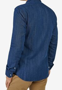 Camicia in denim blu scuro con maniche lunghe, colletto e polsini con bottone; texture liscia con una leggera lucentezza e orlo curvo.