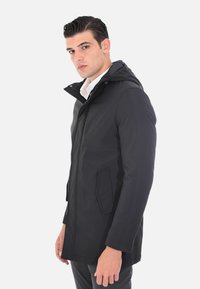 Giacca nera impermeabile con cappuccio, dotata di zip frontale, due tasche laterali e una texture liscia, indossata sopra una camicia bianca.