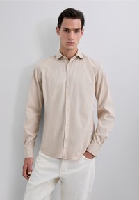 Camisa color beige claro de botones, hecha de algodón suave, con cuello, mangas largas y detalles de costura sutiles. Textura suave y corte regular.