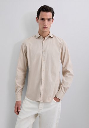 Camisa color beige claro de botones, hecha de algodón suave, con cuello, mangas largas y detalles de costura sutiles. Textura suave y corte regular.